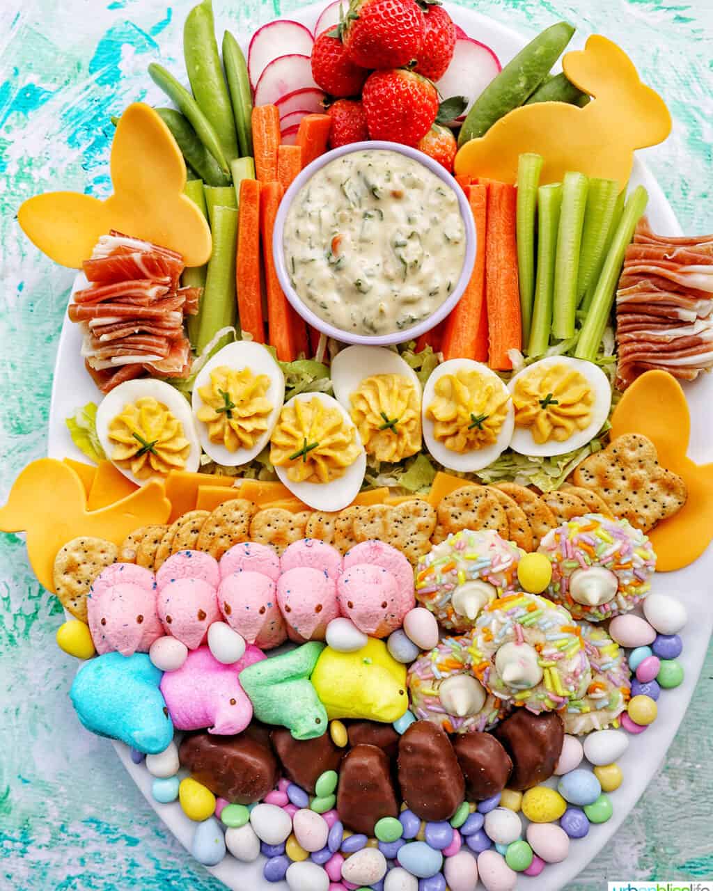 Peeps, chocolate Easter candies, crackers, and deviled eggs on shredded lettuce next to sliced vegetables and a bowl of vegetable dip on a large white platter.