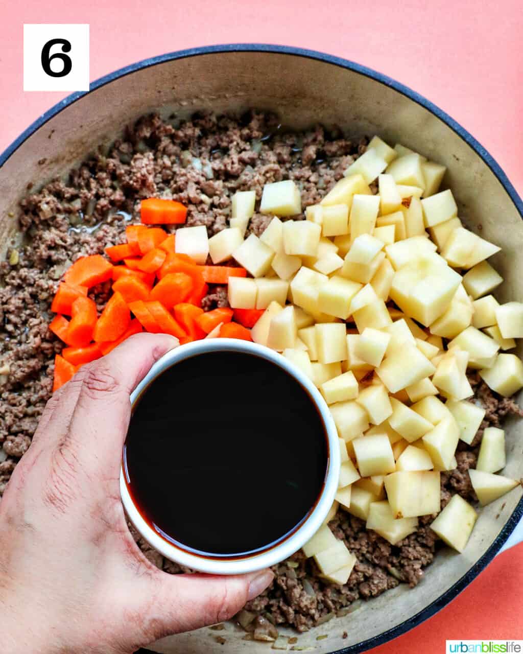 hand holding a bowl of soy sauce mixture over chopped potatoes, carrots, and ground beef in a large pan.