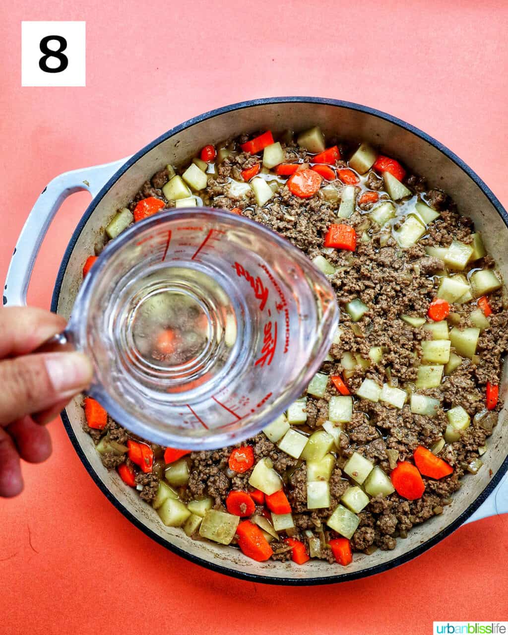 hand holding a measuring cup of water over a large pan of Filipino beef picadillo.