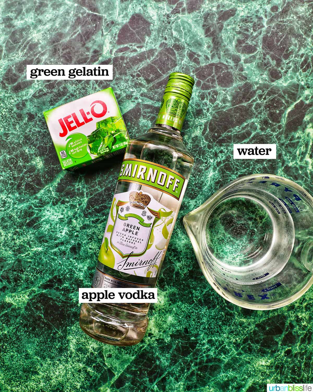 box of jello, bottle of apple vodka, and cup of water on a bright green table.