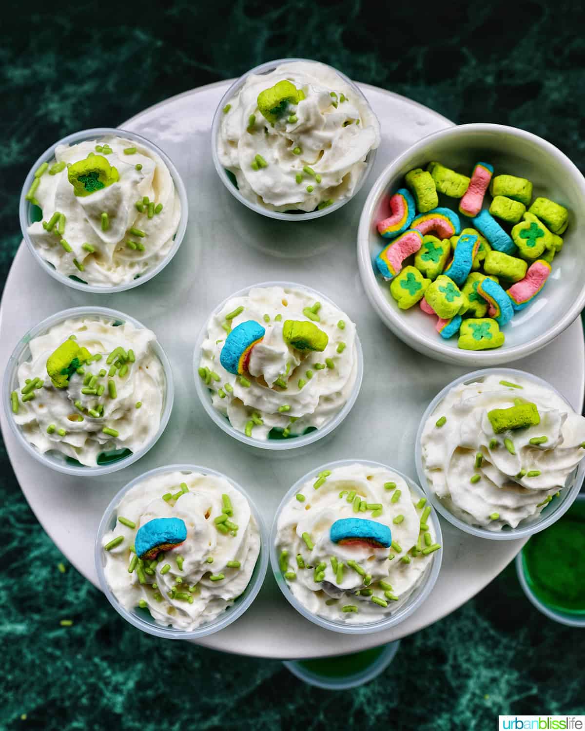 several green jello shots topped with whipped cream and colorful marshmallows on a cake pedestal.