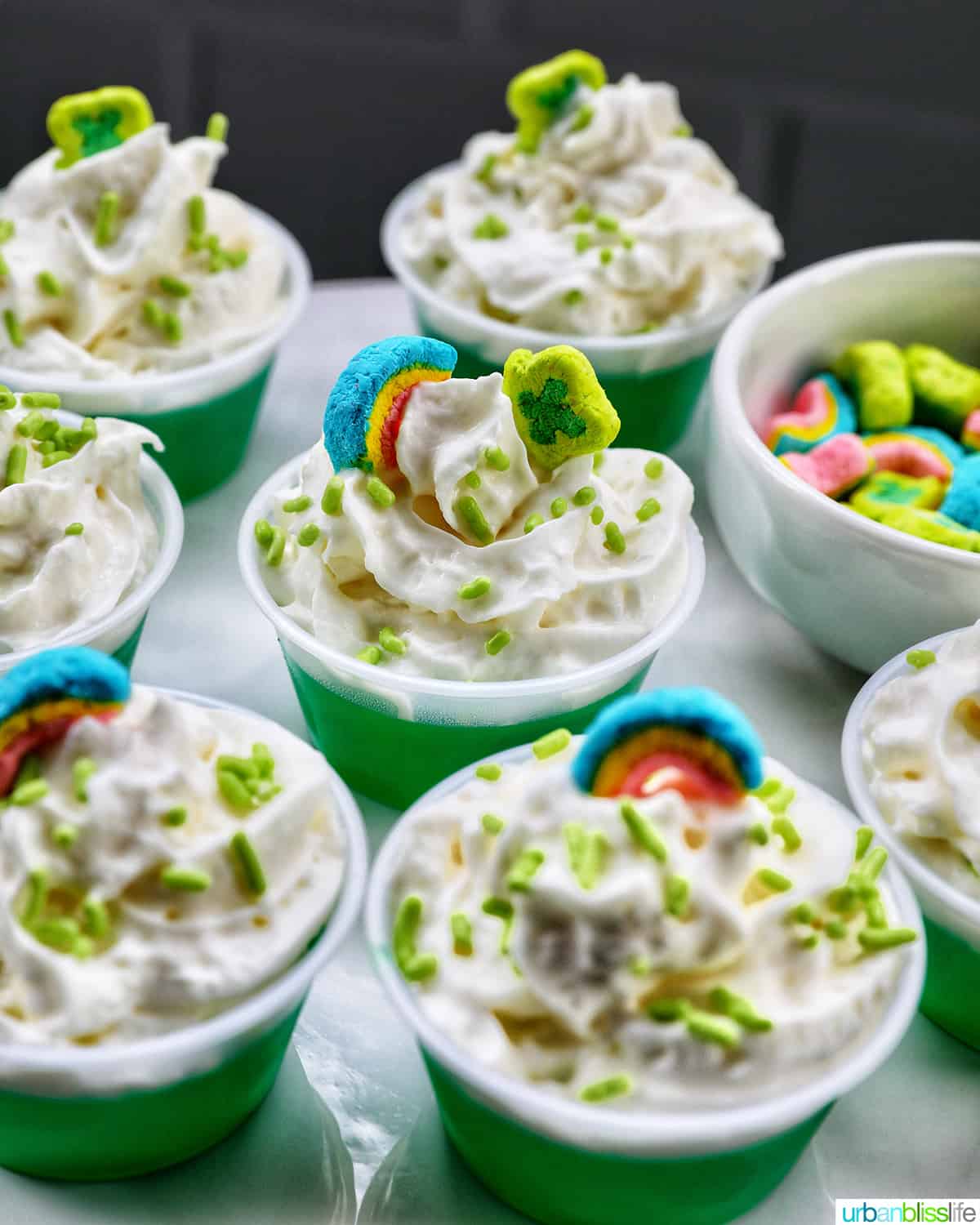 several green jello shots topped with whipped cream and colorful marshmallows on a cake pedestal.