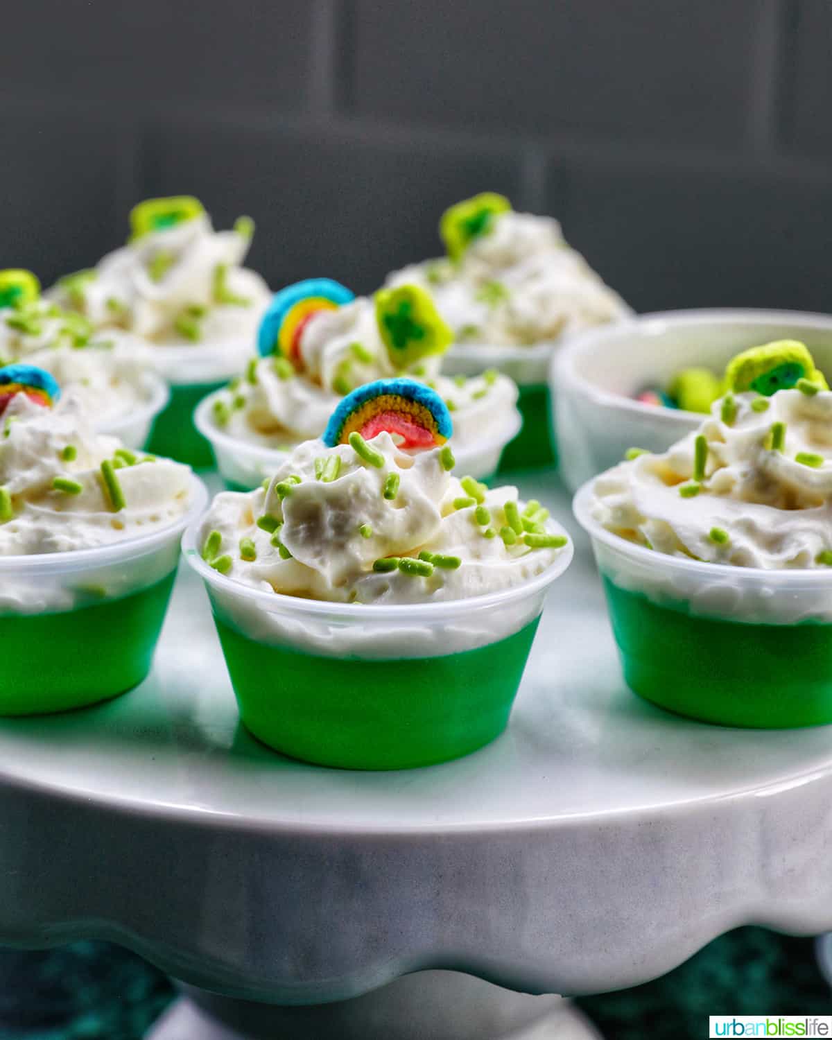 several green jello shots topped with whipped cream and colorful marshmallows on a cake pedestal.