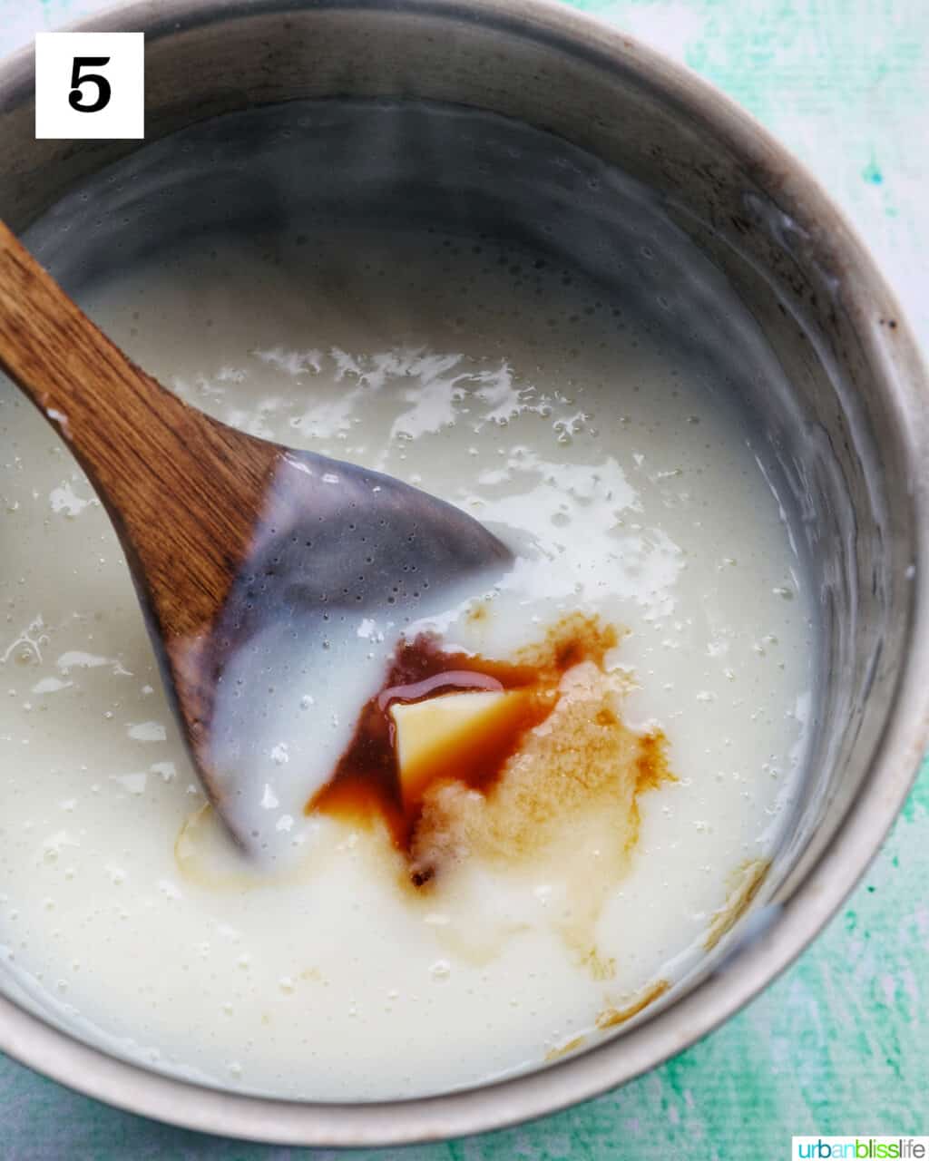 wooden spoon stirring vanilla extract and butter into a saucepan of vanilla pudding.