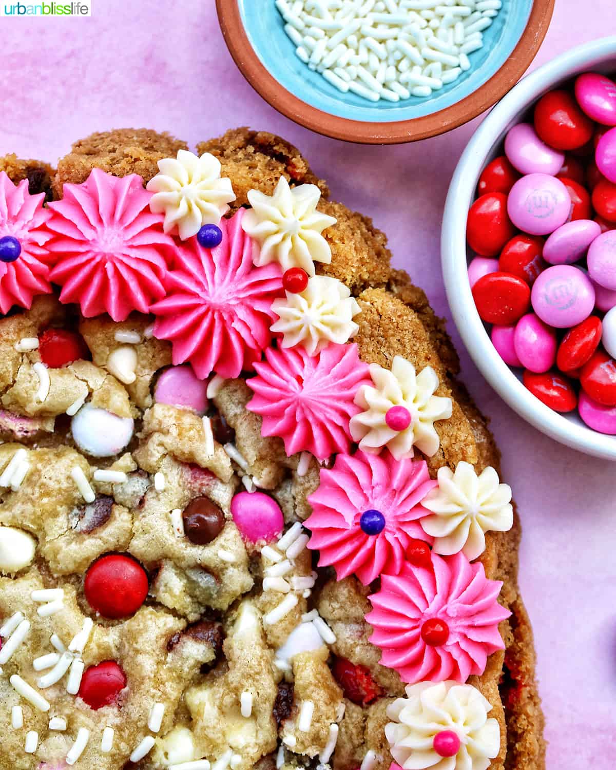 A heart-shaped Valentine's Day cookie cake with pink and white vanilla frosting and red, white, and pink M & M decorations.