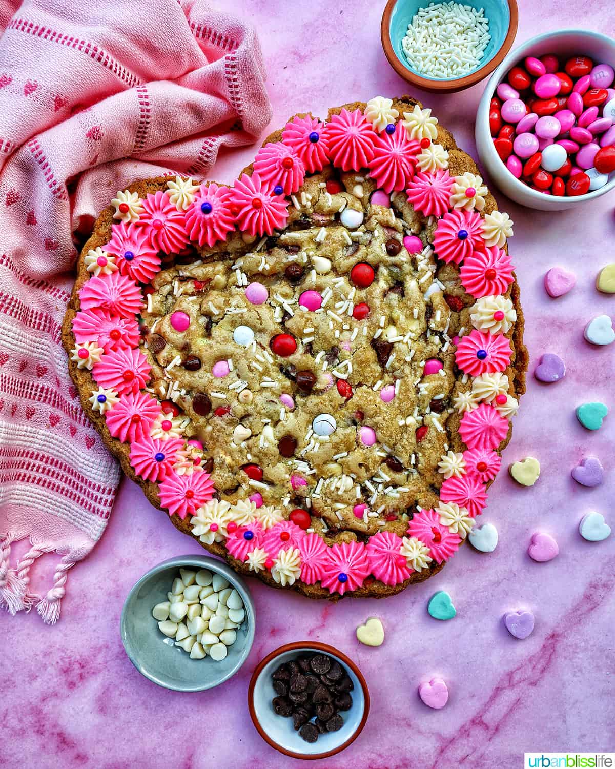 A Valentine's Day Cookie Cake with pink frosting, pink, red and white M & M candies, conversation hearts candies sprinkled around the cookie cake, and white sprinkles.