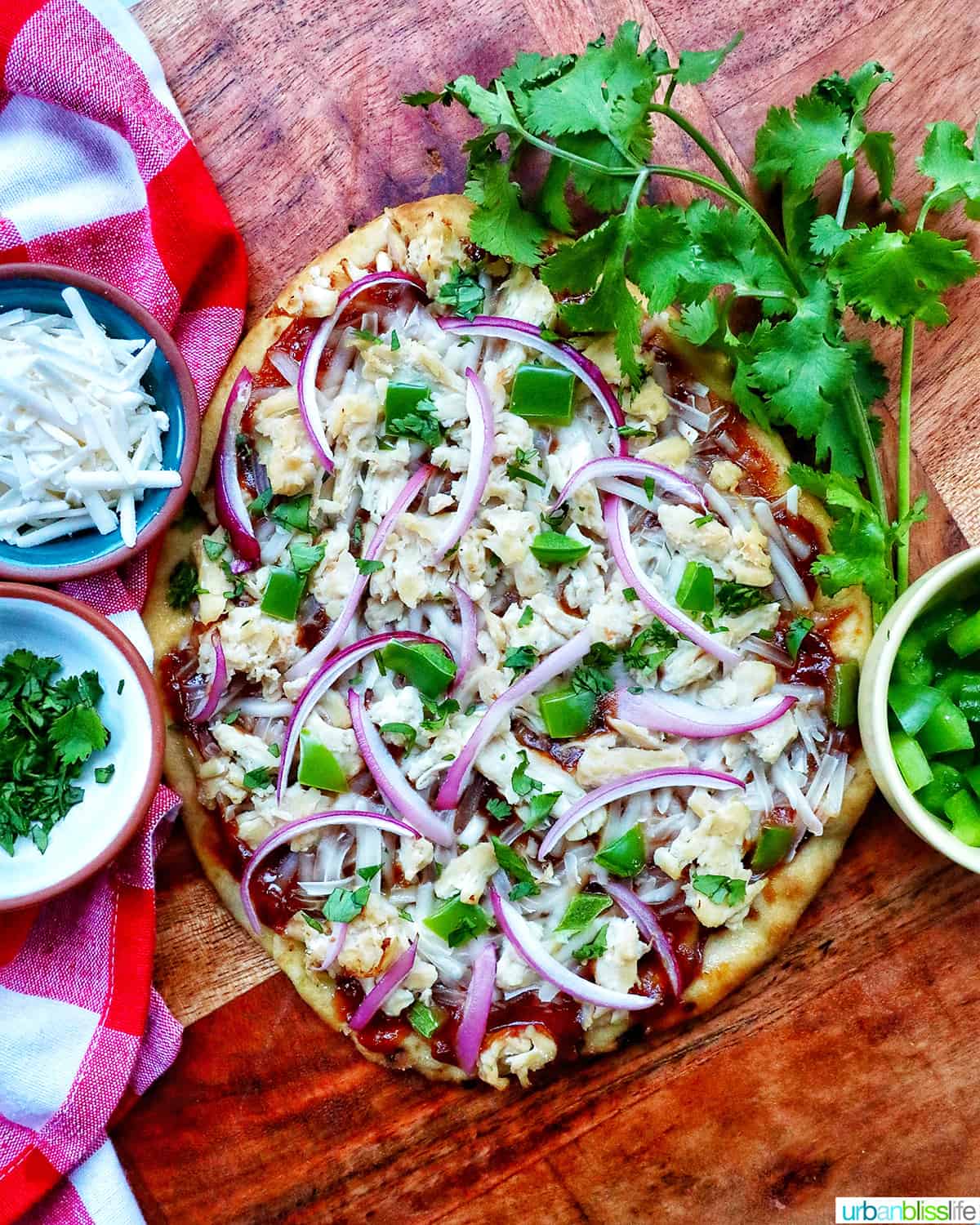 barbecue chicken flatbread pizza with red onions, chicken breast, green peppers, and cilantro on a wooden board with sides of cilantro leaves and shredded cheese.