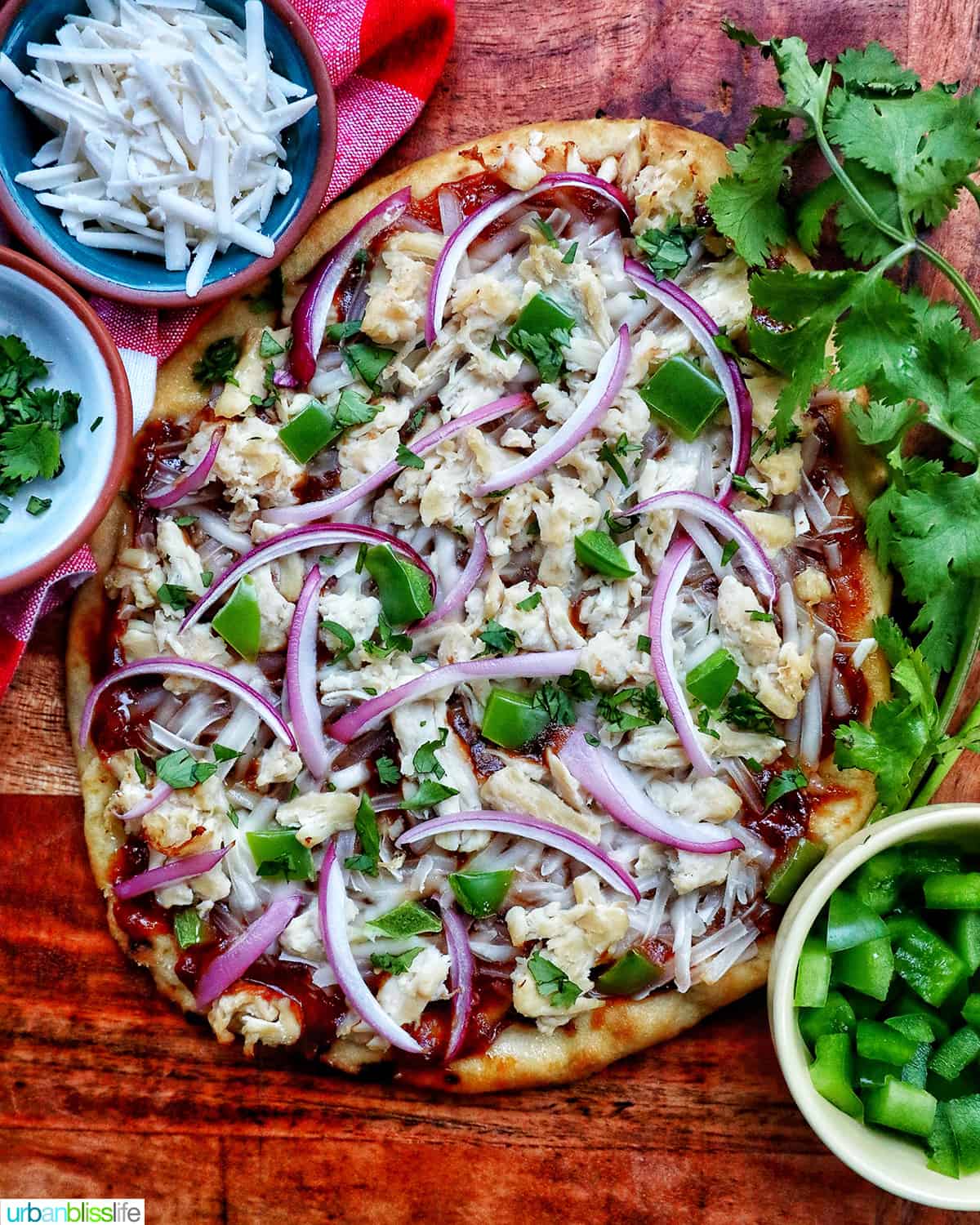 barbecue chicken flatbread pizza with red onions, chicken breast, green peppers, and cilantro on a wooden board with sides of cilantro leaves and shredded cheese.