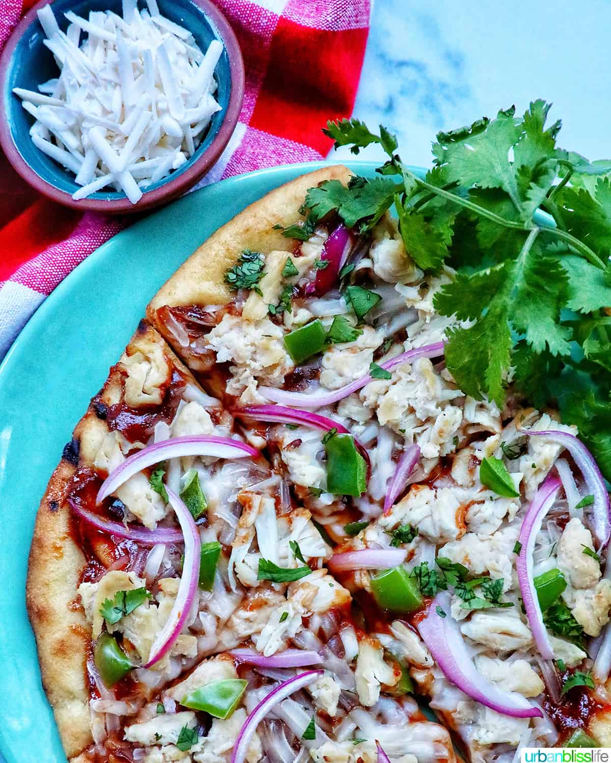 barbecue chicken flatbread pizza with red onions, chicken breast, green peppers, and cilantro on a wooden board with sides of cilantro leaves and shredded cheese.