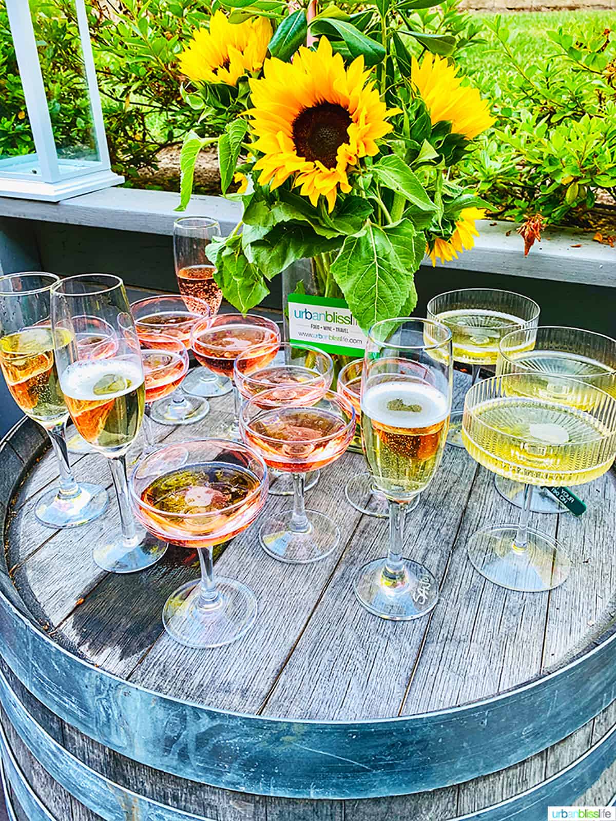 several glasses of rosรฉ wine and Champagne on a wine barrel with vase of sunflowers.