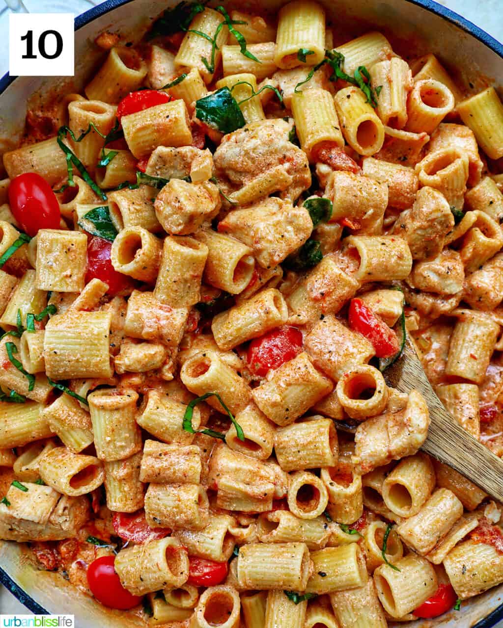wooden spoon in a large skillet of rigatoni pasta in a creamy Tuscan sauce with chicken, tomatoes, and herbs.