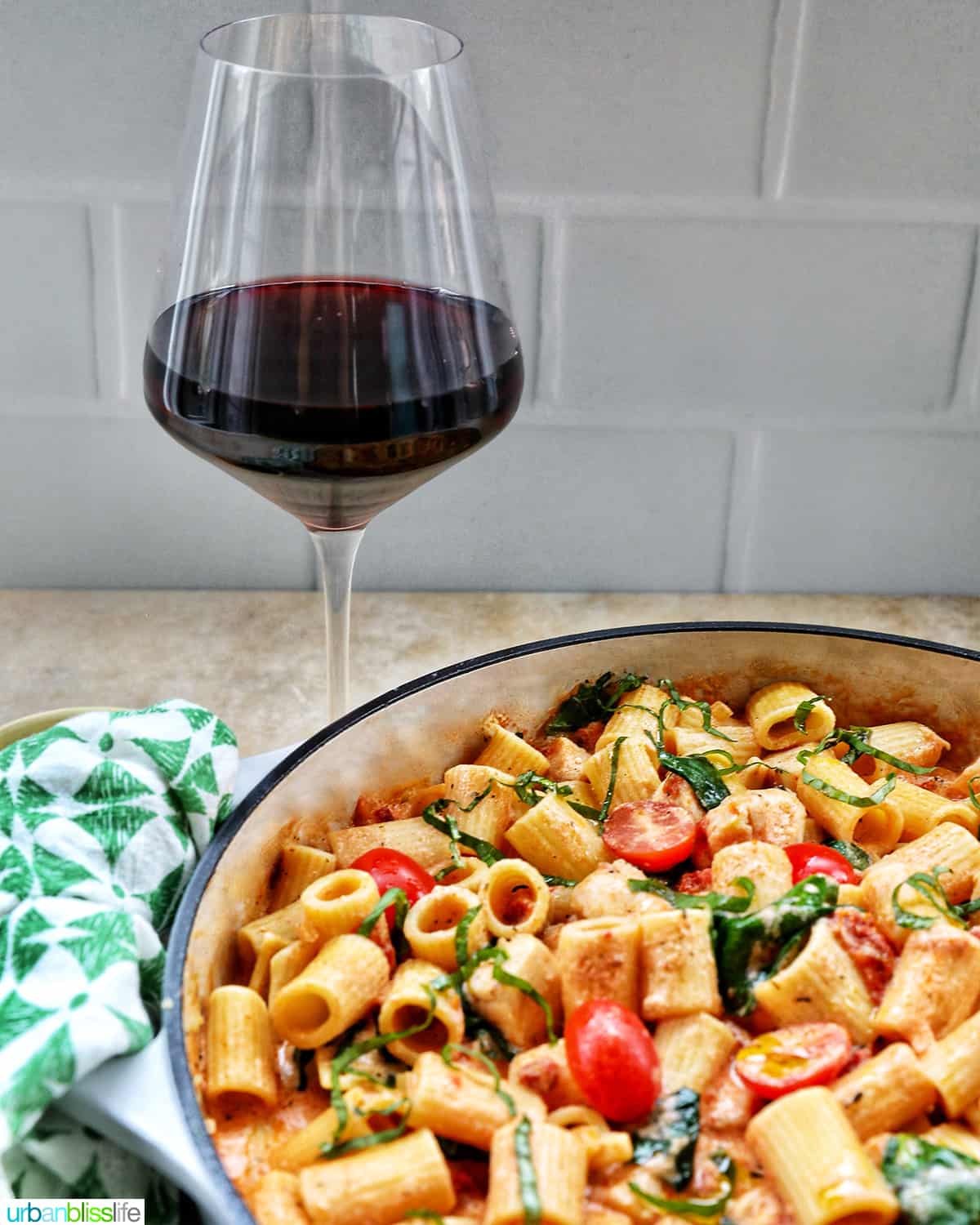 glass of red wine next to a large skillet of rigatoni pasta in a creamy Tuscan sauce with chicken, tomatoes, and herbs.