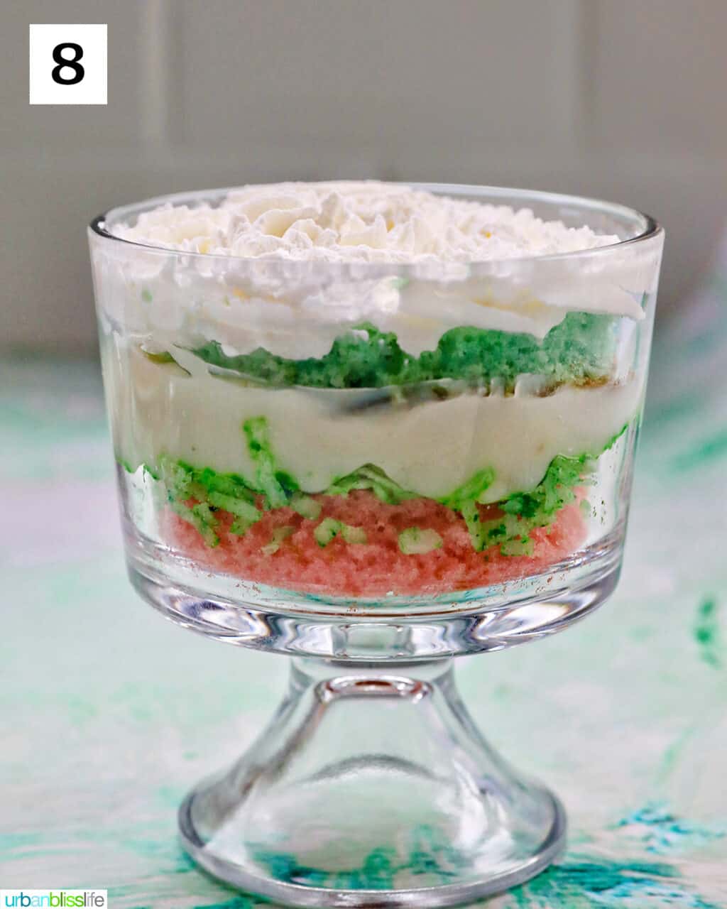 layer of whipped topping over layer of green cake over vanilla pudding over shredded coconut over a layer of pink cake in a trifle bowl.