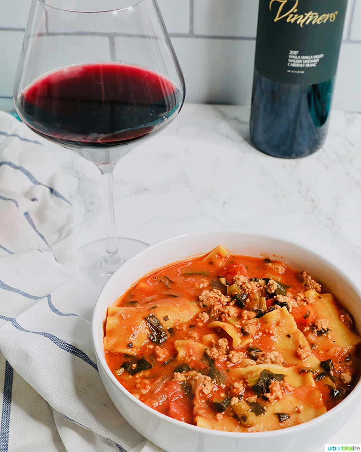 glass of red wine next to a white bowl full of lasagna soup.