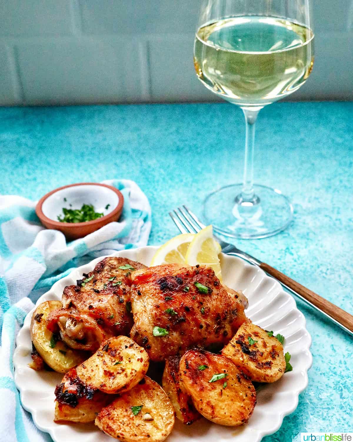 plate of chicken and potatoes next to a fork, glass of white wine, and side of chopped parsley.