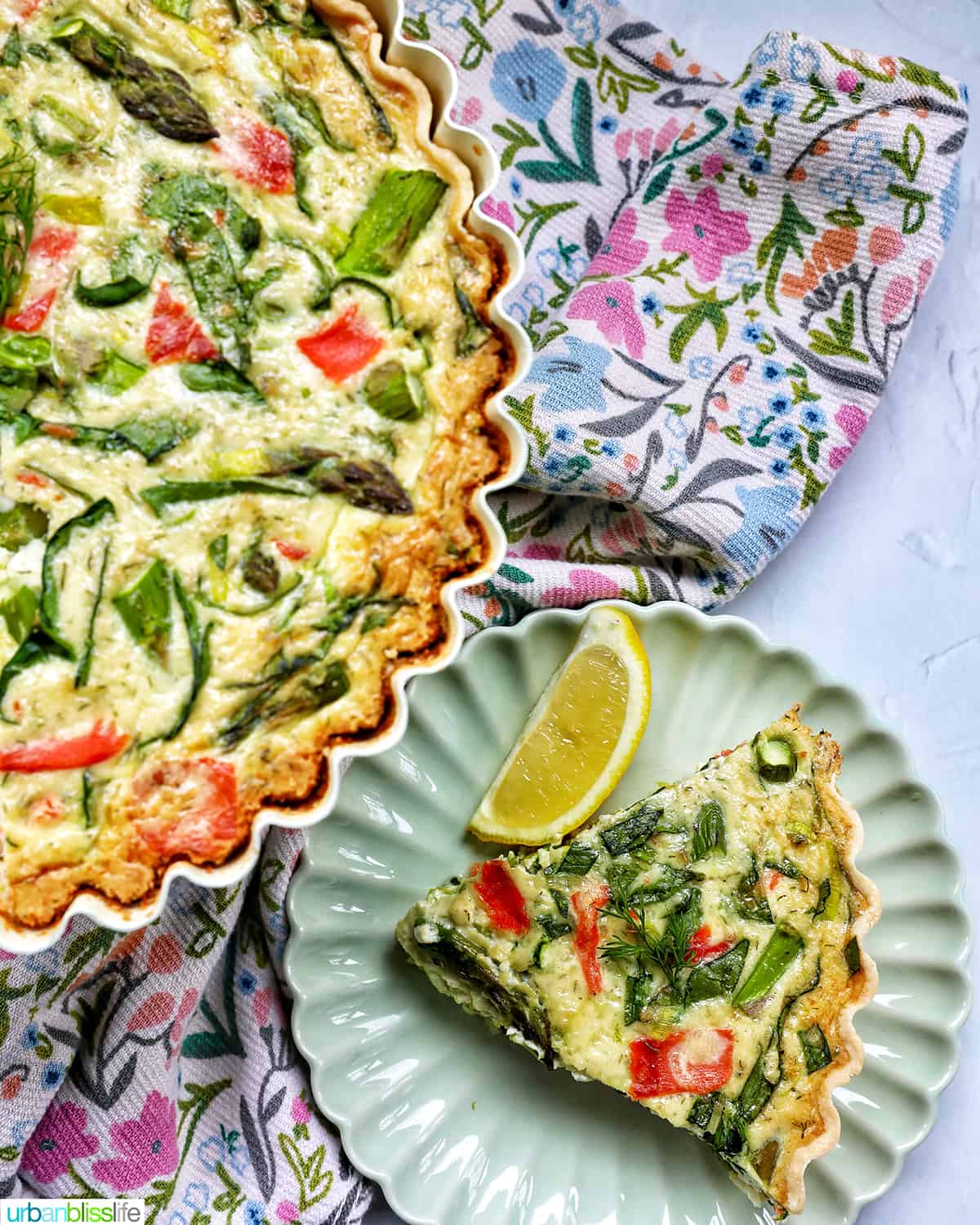 slice of salmon quiche on a plate with a half of a full salmon quiche on a pedestal and floral napkin.