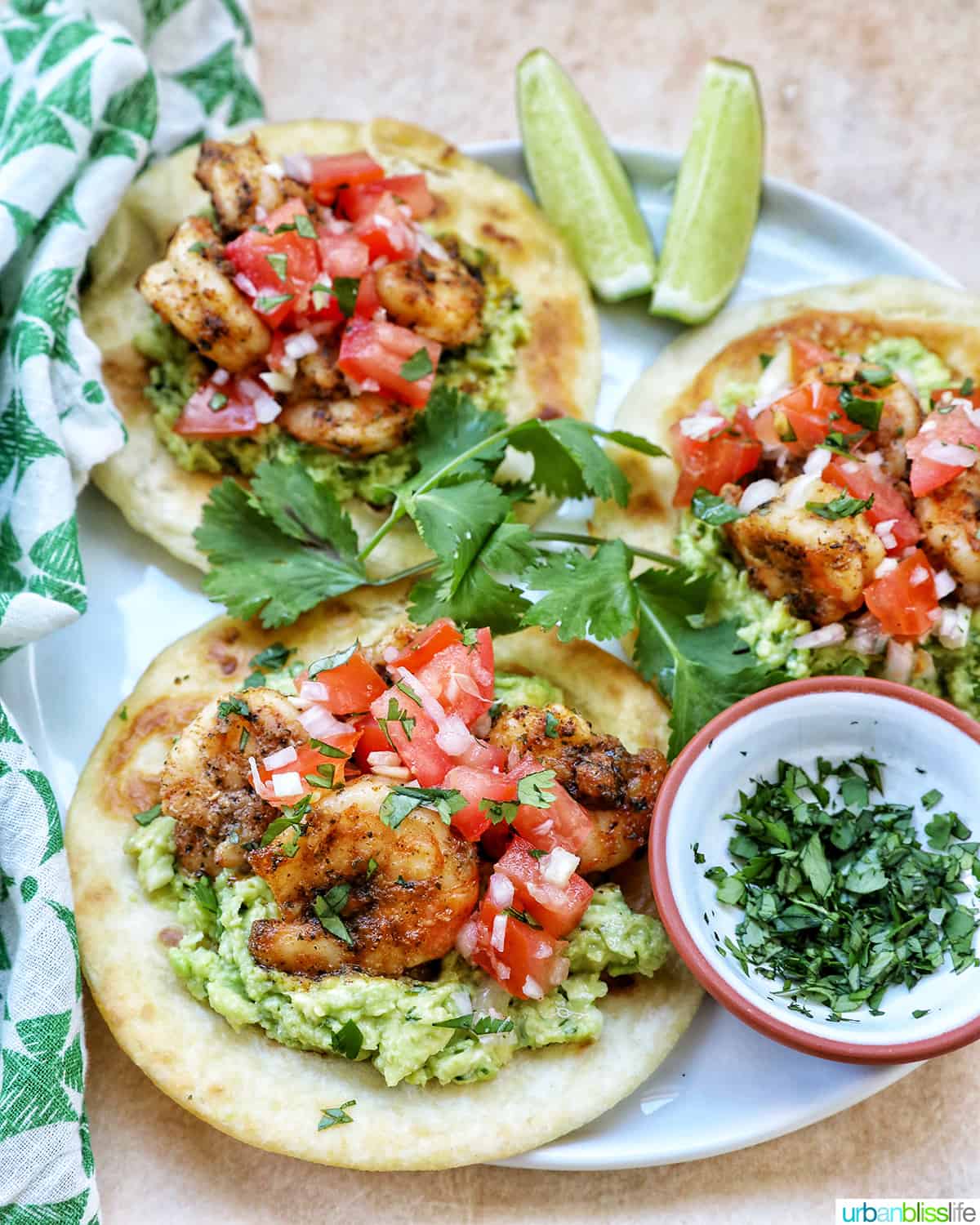 three shrimp tostafas with pico de gallo, shrimp, and guacamole on a tortilla with cilantro leaves on the side.