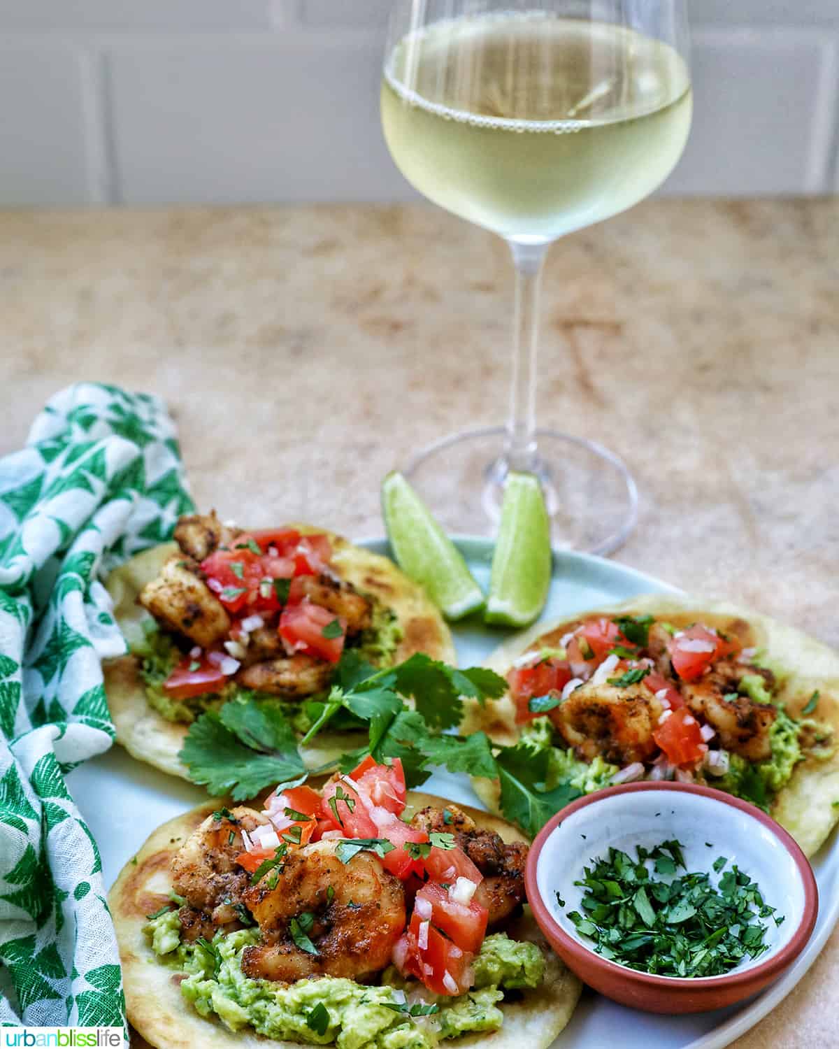 three shrimp tostafas with pico de gallo, shrimp, and guacamole on a tortilla with cilantro leaves on the side and a glass of white wine.