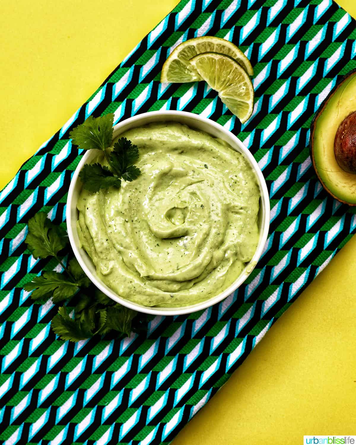 bowl of avocado cilantro lime crema on a colorful modern napkin with garnish of cilantro leaves and side of sliced avocado.