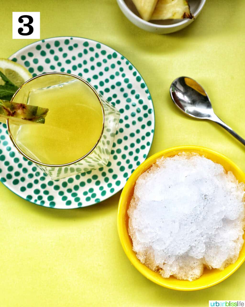 glass of pineapple juice next to a bowl of crushed ice and a spoon.