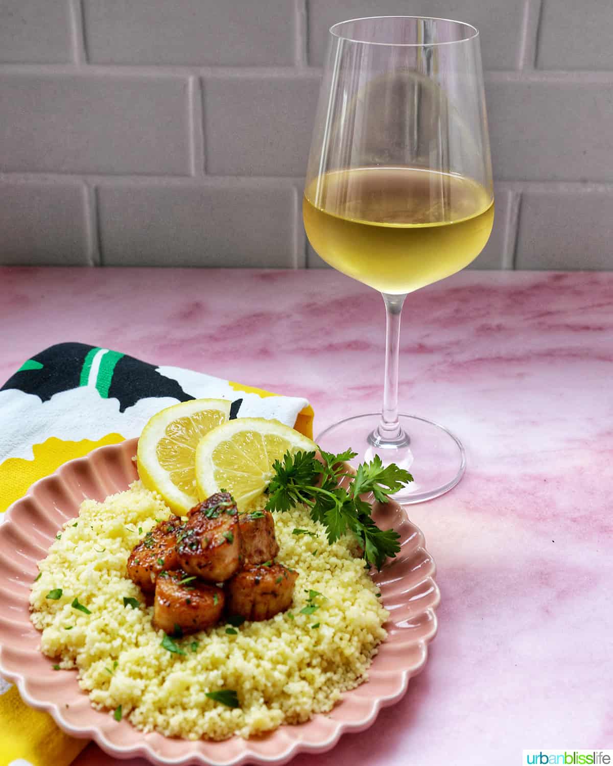 cast iron seared scallops on a bed of couscous garnished with herbs and lemon slices and a glass of white wine.