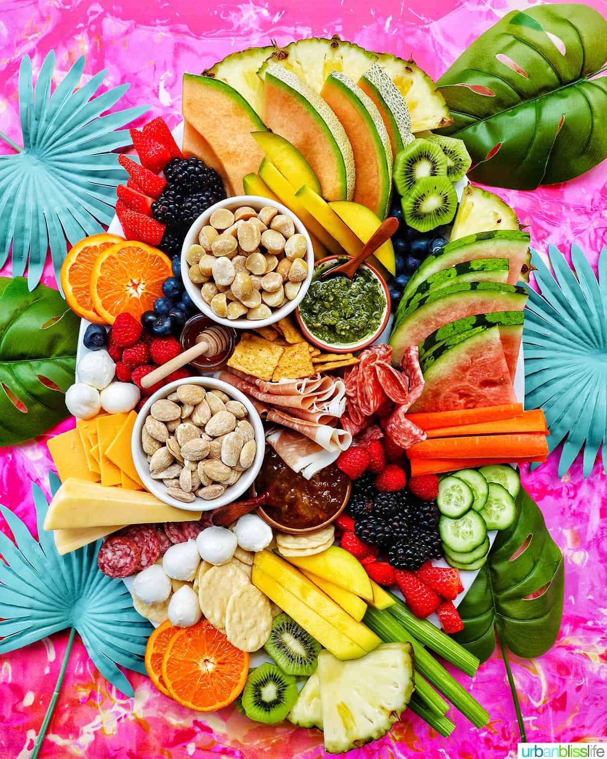Summer charcuterie board with various fruits, vegetables, crackers, meats, and cheeses on a bright pink background.