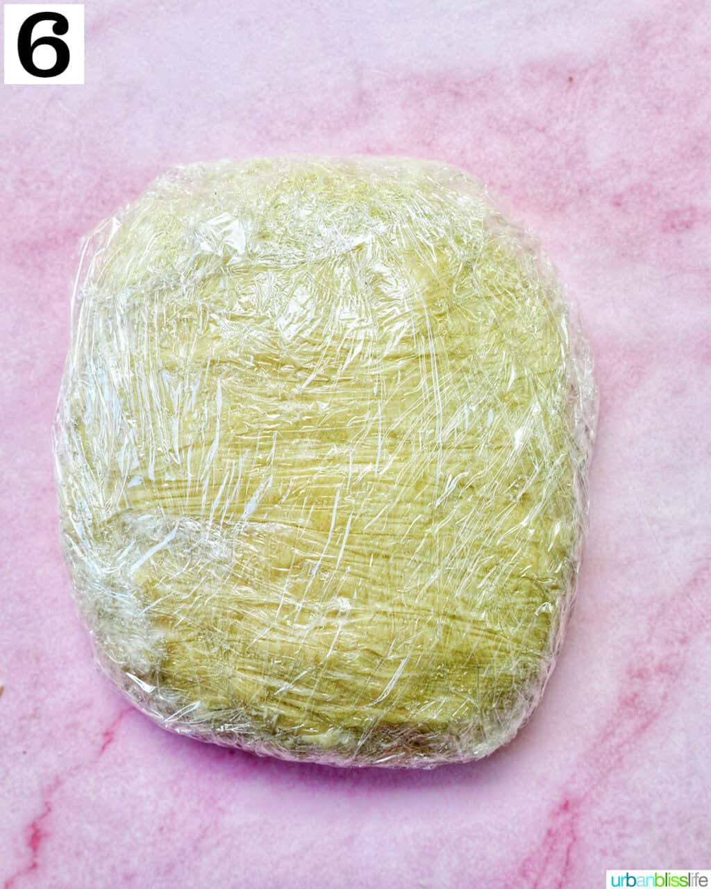 homemade pie dough wrapped in plastic wrap on a pink background.