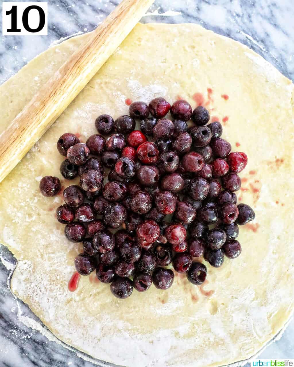 cherry filling in the center of rolled out pie dough on a marble countertop with rolling pin.