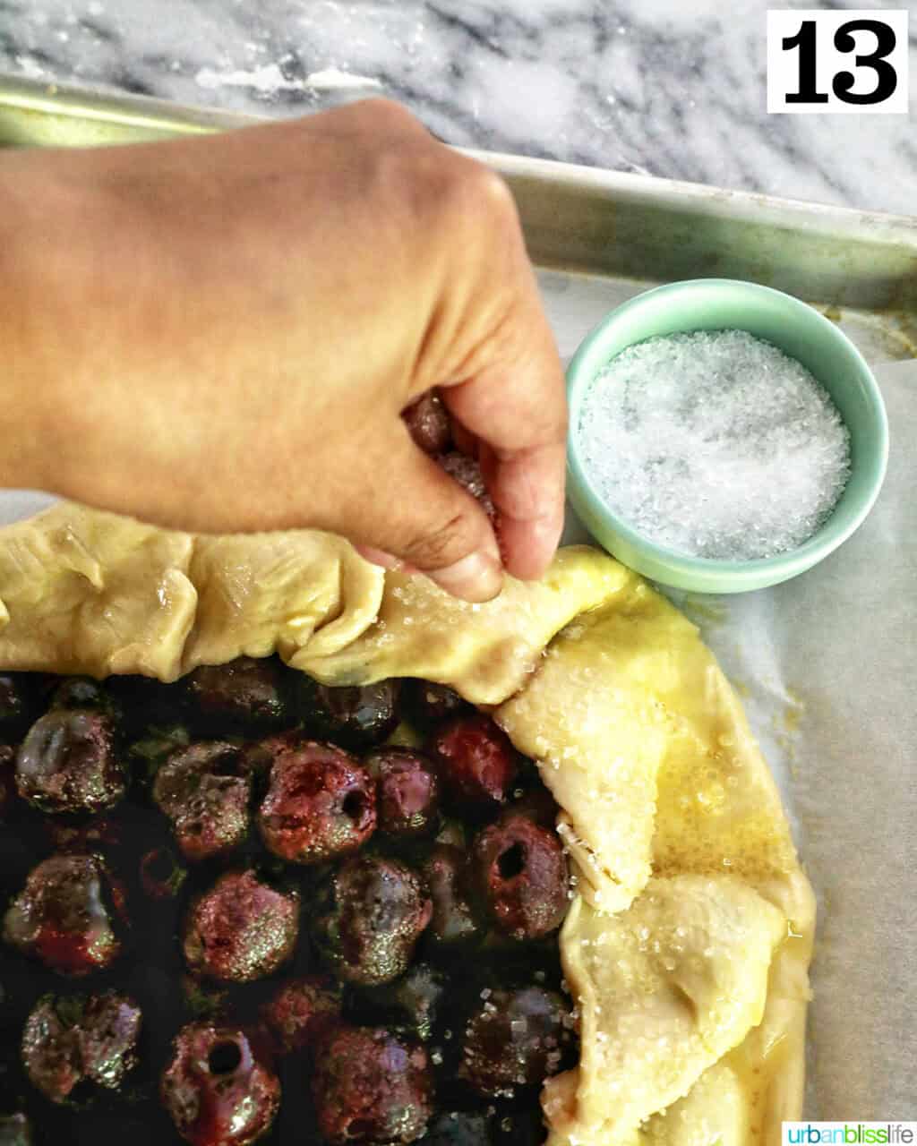 hand sprinkling sanding sugar over the top of the edges of a cherry galette.