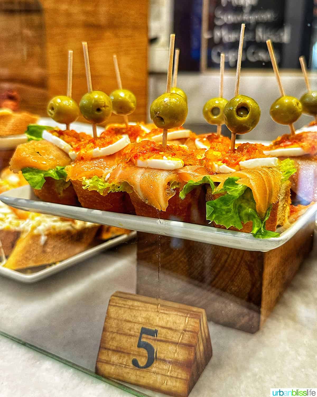 pintxos on a plate pedestal with the number 5 on a wood board in front of it.