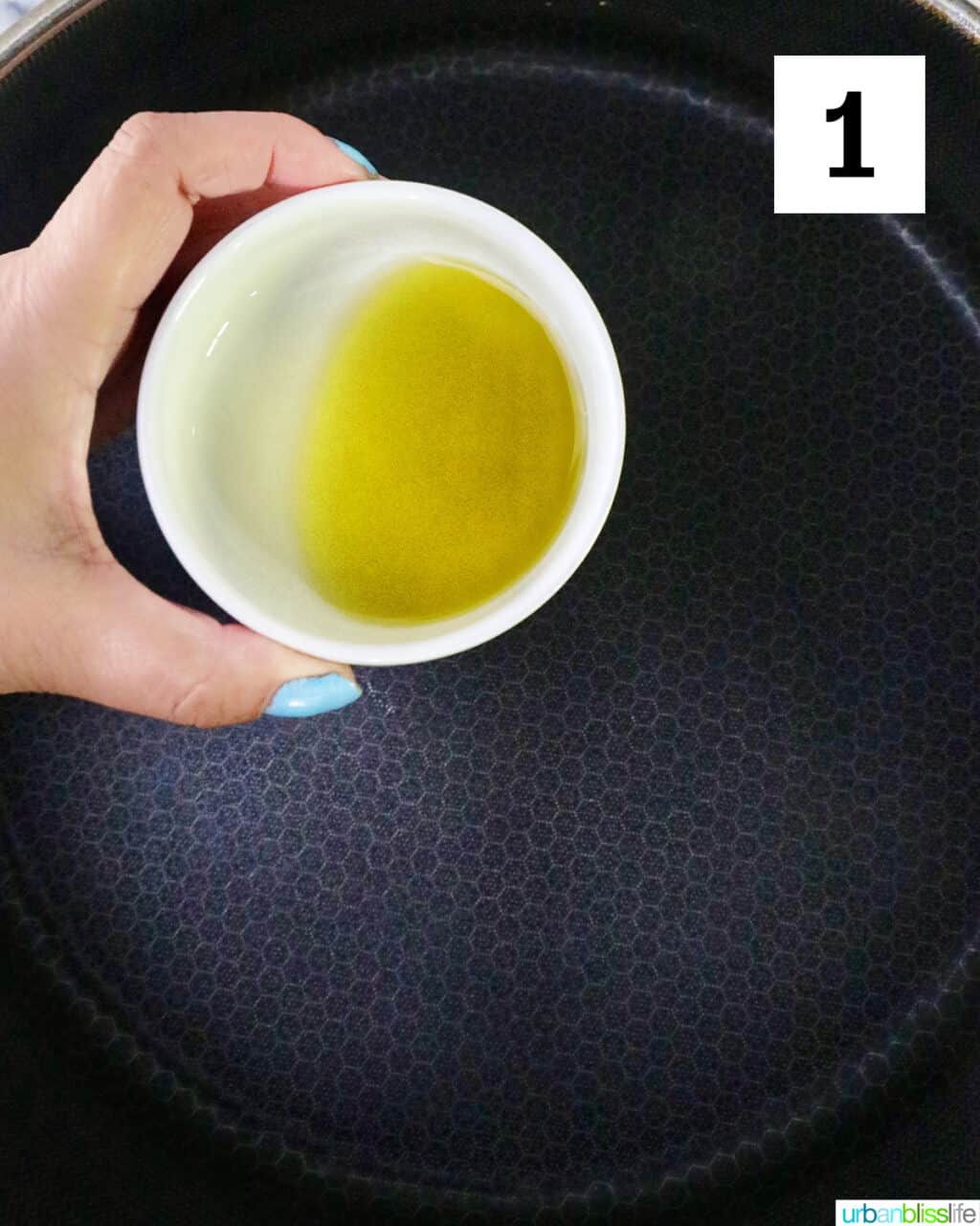 hand holding a bowl of olive oil over a nonstick skillet.