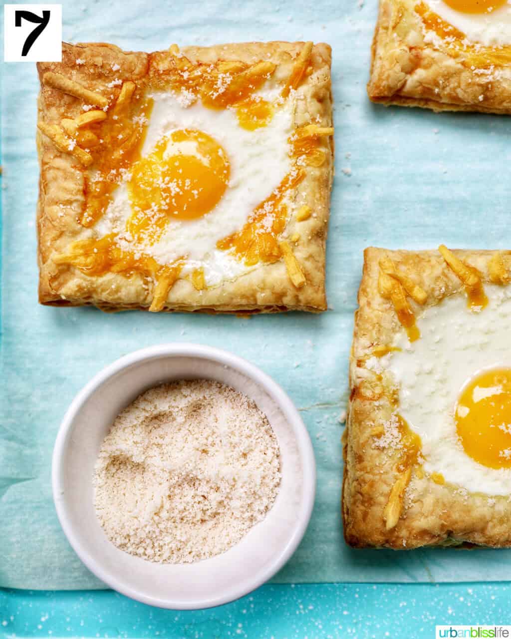 eggs baked in enter of puff pastry squares on a baking sheet with bowl of grated parmesan cheese.