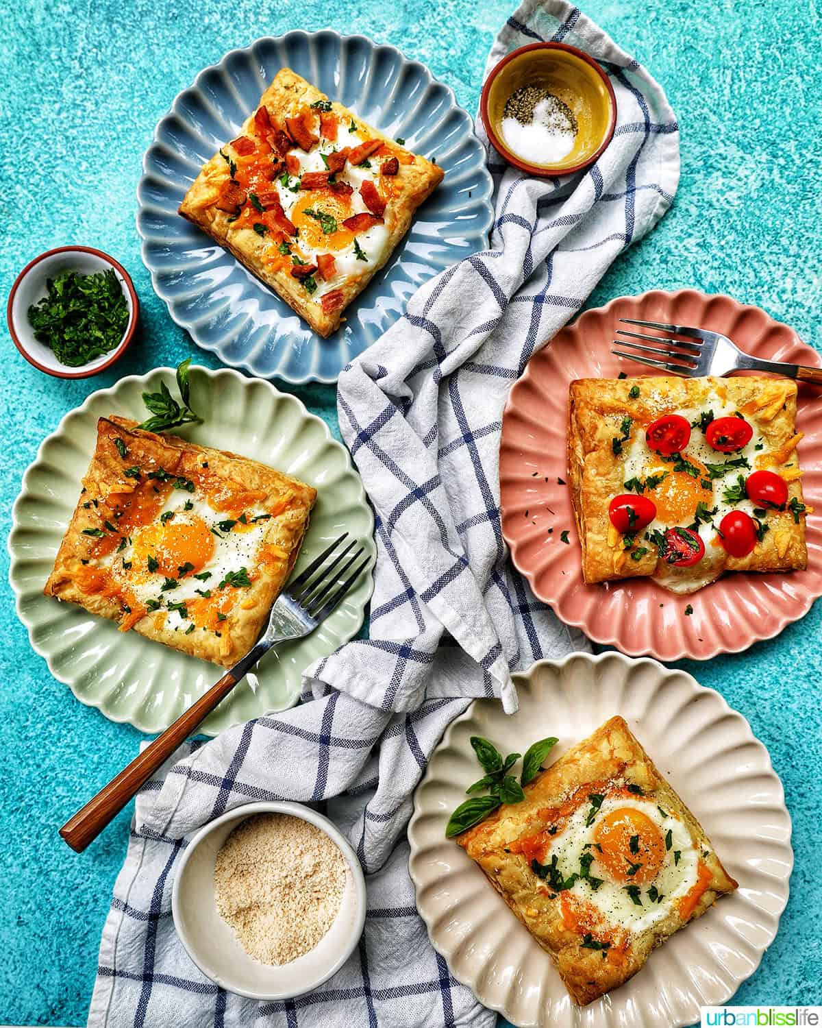 several plates with eggs in puff pastry, one with bacon, one with tomatoes, and a napkin, forks, and parsley garnish.