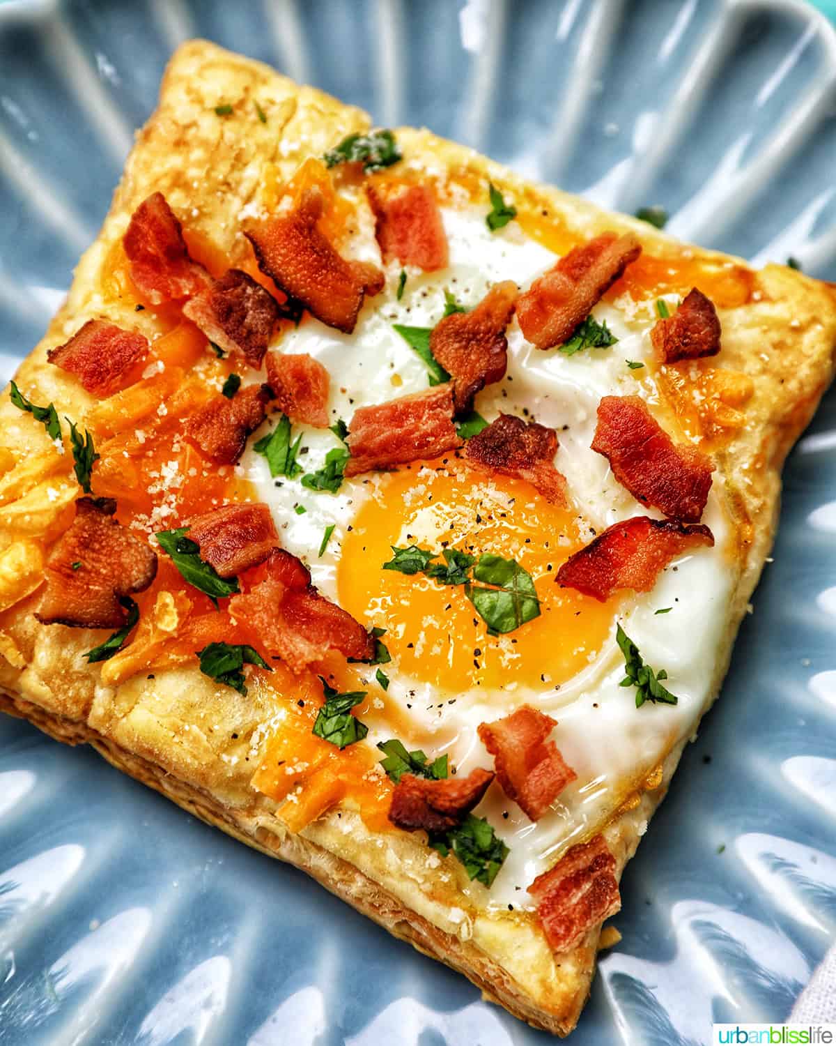 egg in puff pastry on a plate with bacon crumbles and parsley garnish.