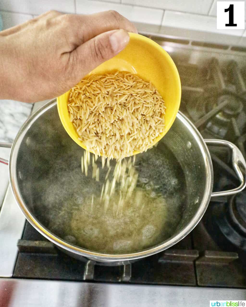 hand holding a yellow bowl pouring orzo pasta into a pot of boiling water.
