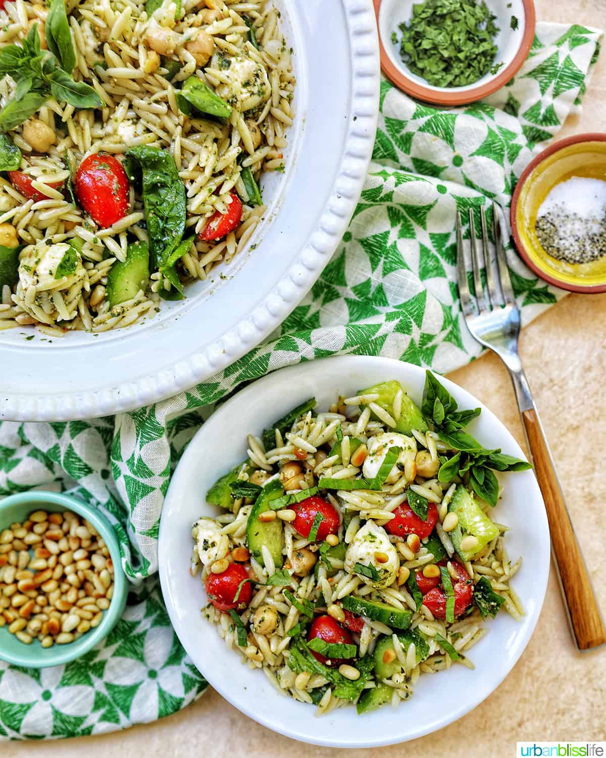 large bowl and small bowl of orzo pesto pasta salad with a fork and small bowls of garnishes.