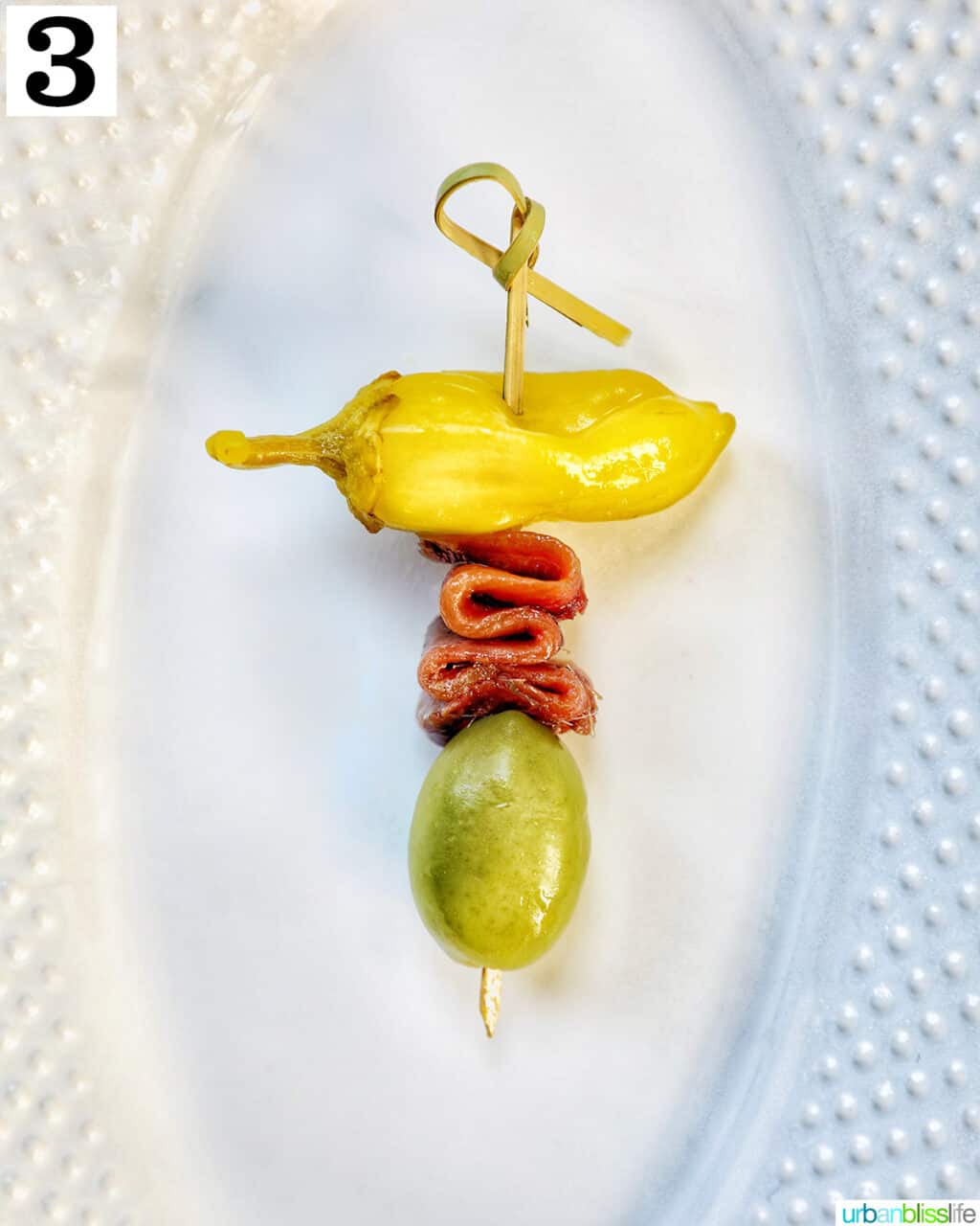 white plate with a single food pick with pepper, anchovy, and olive to create a Gilda pintxo.