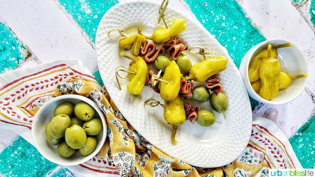 Gilda pintxos: food sticks with peppers, anchovies, and olives on a white plate with bowls of olives next to it.