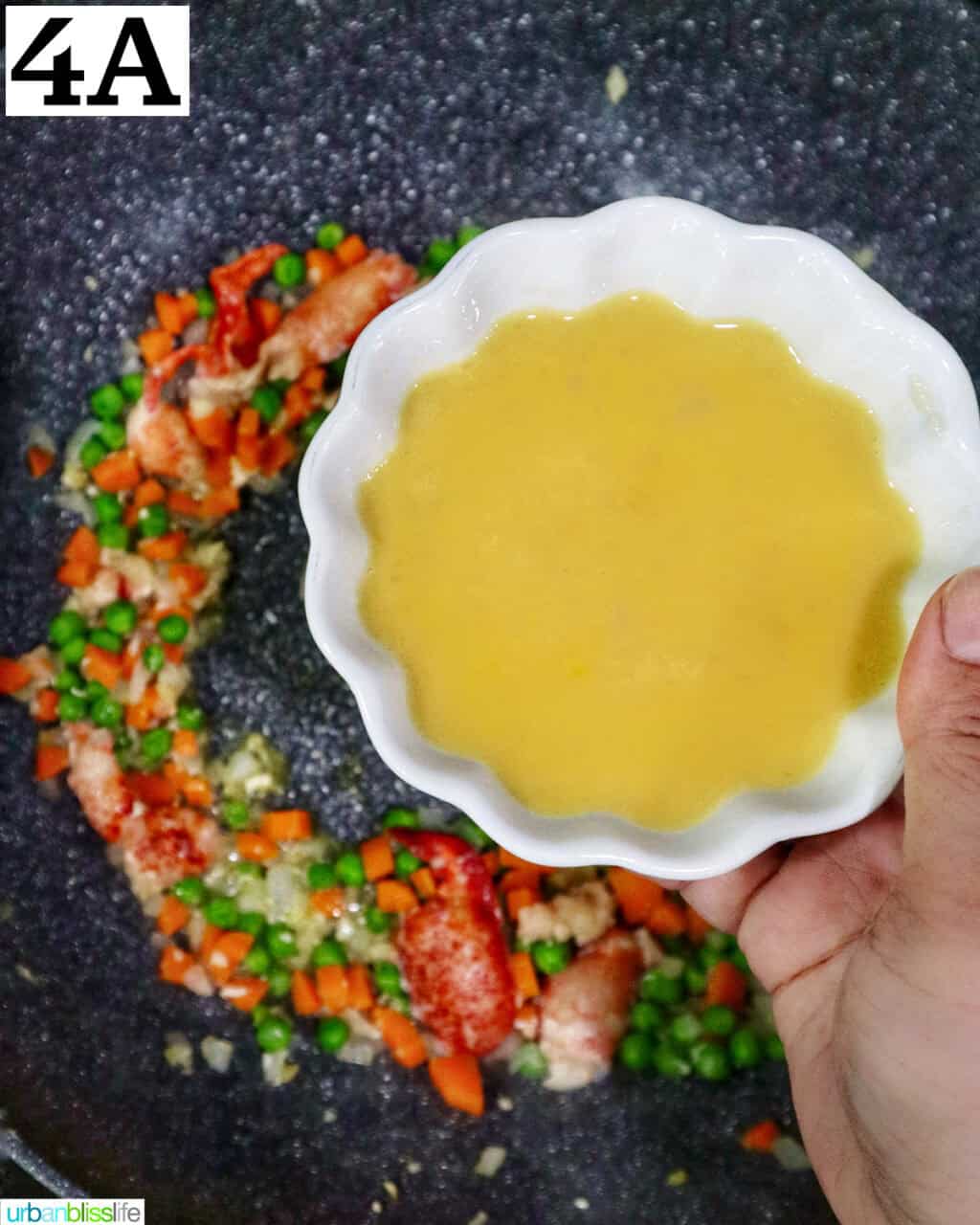 hand holding a bowl of beaten eggs over a wok with fried rice.