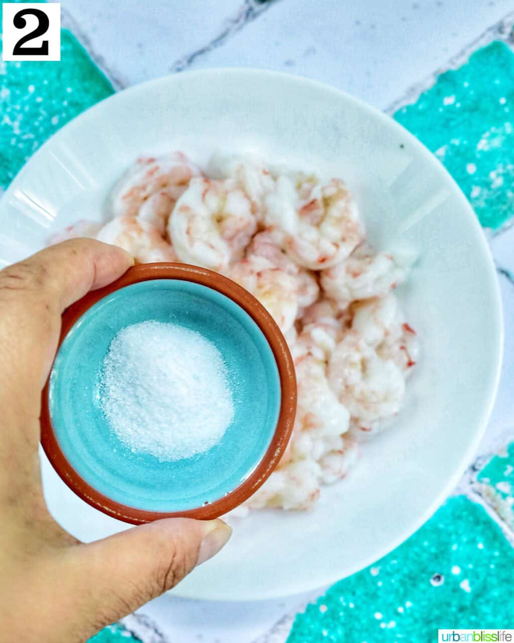 hand holding a small bowl of salt over a bowl of shrimp.