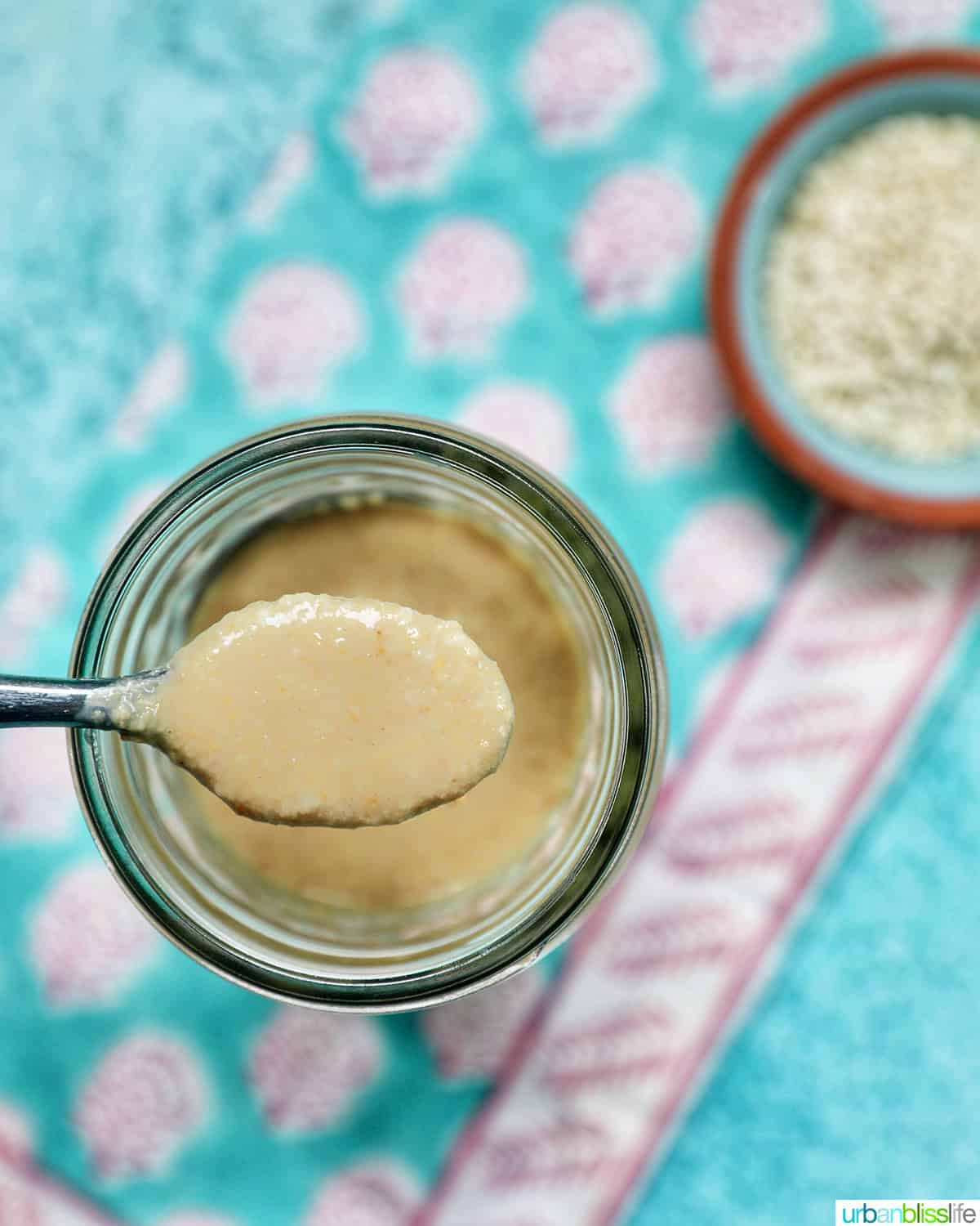 jar of homemade tahini with a small bowl of sesame seeds and a spoon.