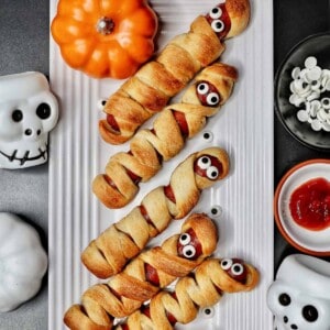 mummy hot dogs on a white plate surrounded by Halloween decorations like mini pumpkins and skull bowls.