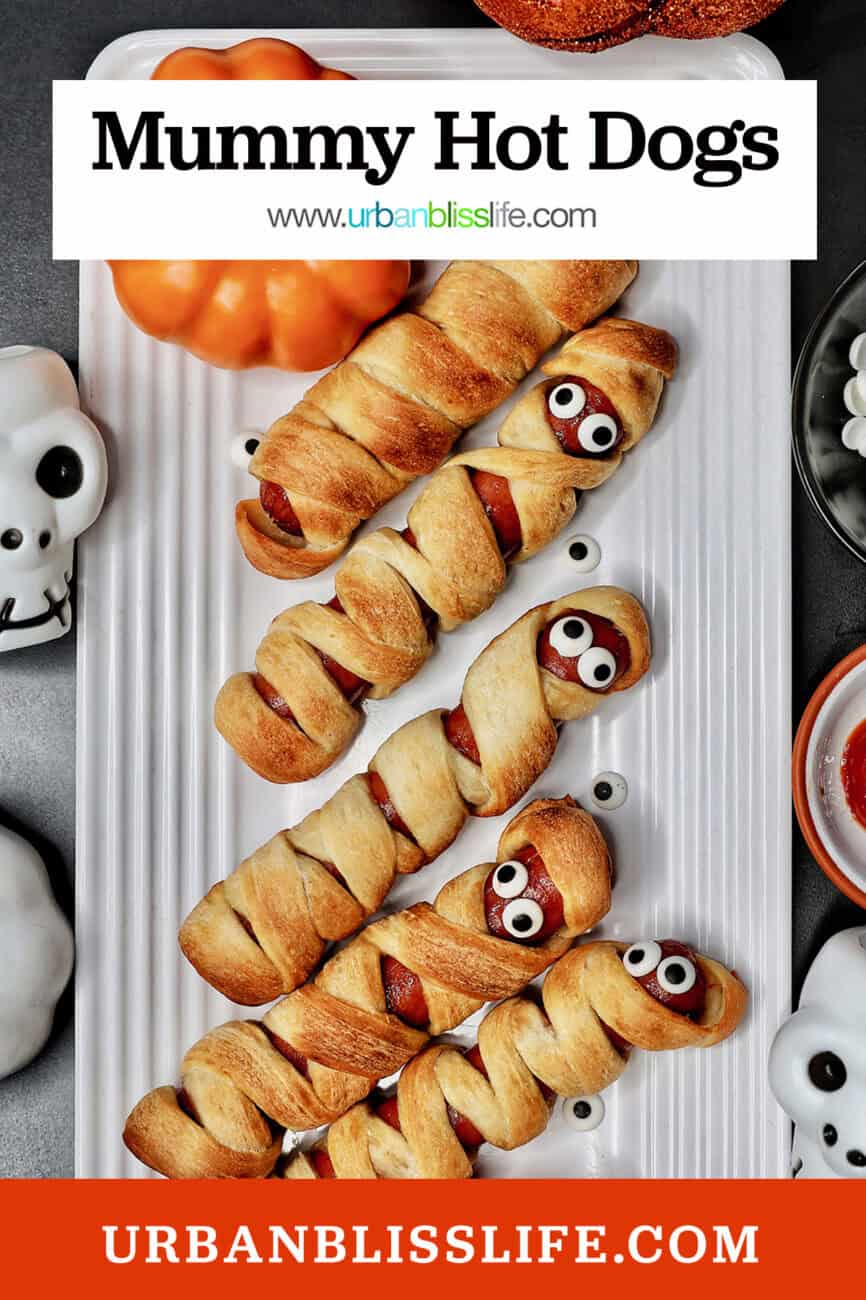 mummy dogs - hot dogs wrapped in pizza dough - stacked on a white plate next to Halloween decorations with title text overlay.