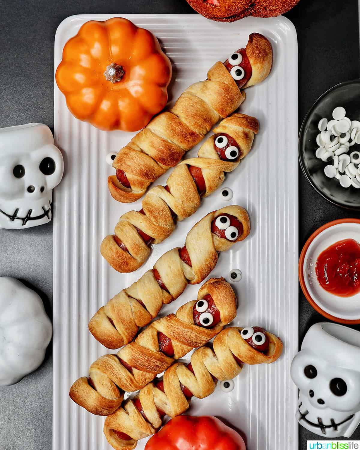 mummy hot dogs on a white plate surrounded by Halloween decorations like mini pumpkins and skull bowls.