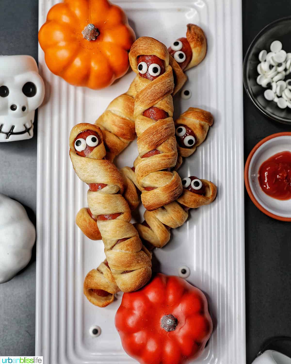mummy hot dogs on a white plate surrounded by Halloween decorations like mini pumpkins and skull bowls.