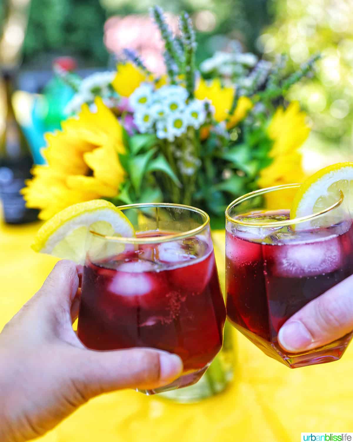 hands holding glasses of glass of tinto de verano wine cocktail with lemon slice by summer flowers.