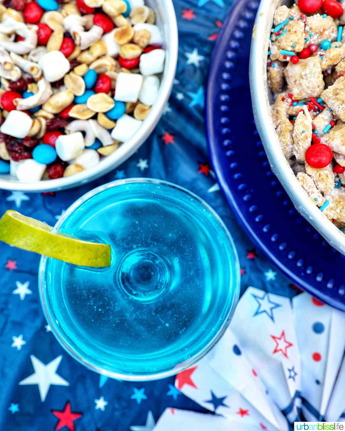 overhead of a blue wave cocktail with bowls of red, white, and blue puppy chow and trail mix.
