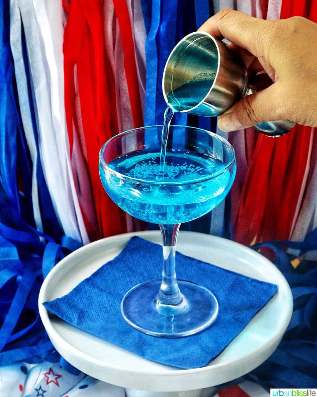 pouring curacao into a cocktail glass with red, white, and blue decorations in the background.
