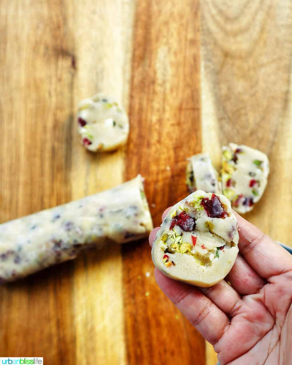hand holding a sliced o make pistachio cranberry shortbread cookie dough over a cutting board.