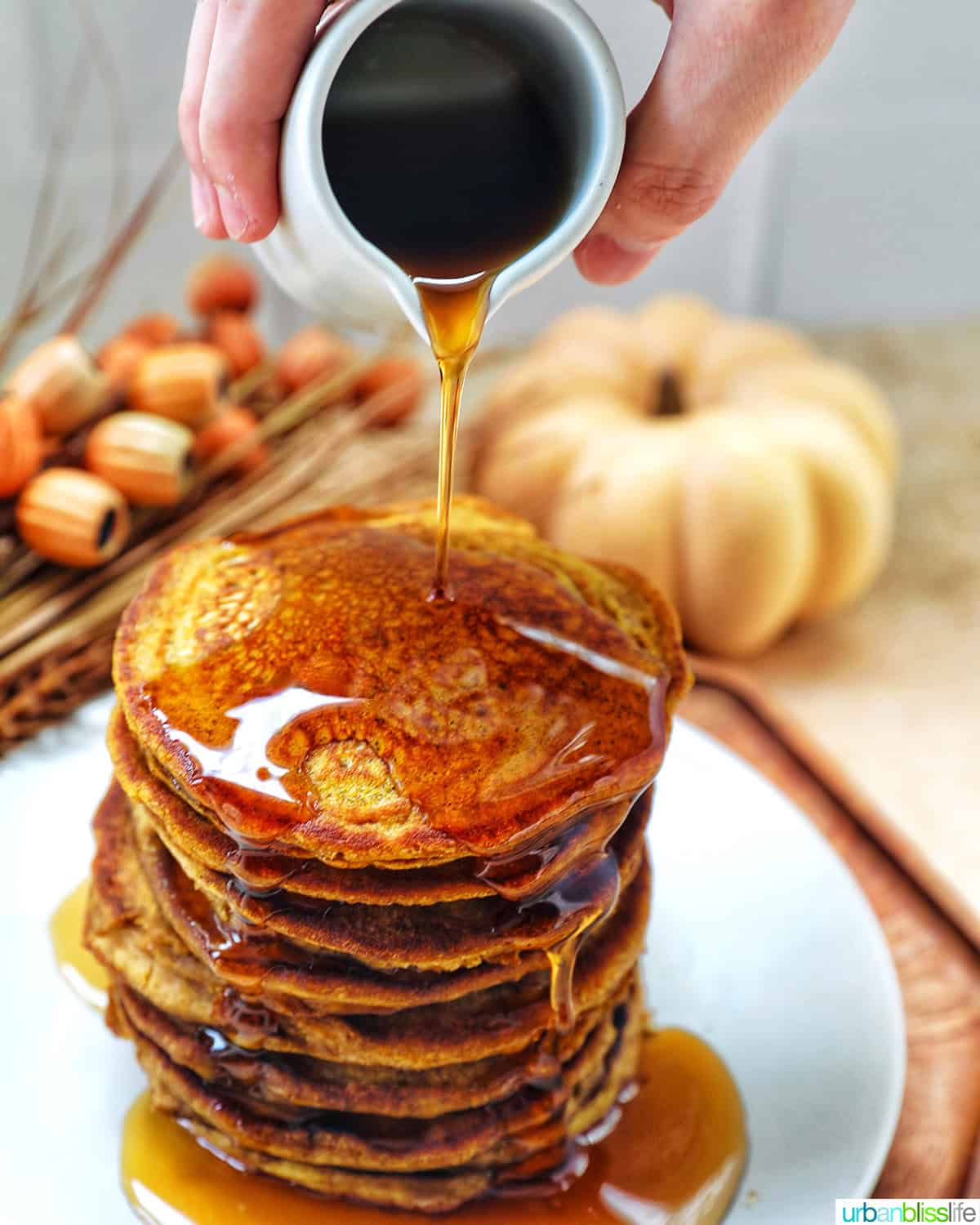 stack of pumpkin spice pancakes on a plate with hand pouring syrup and fall decorations.